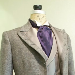 Puede incluir: Chaqueta de traje de tweed gris con chaleco a juego y corbata morada. El traje se exhibe en un maniquí. La chaqueta tiene solapas con muescas y botones. El chaleco es de color gris claro.