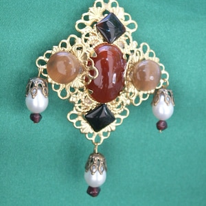 Op de afbeelding: Een gouden ketting met een grote, sierlijke hanger in filigraan. De hanger is bezet met rode, bruine en zwarte edelstenen. De ketting is geregen met witte parels.