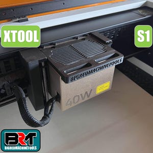 May include: A close-up of a laser machine component with the text "XTOOL" in a green box. The component is black and tan with the text "BIGREDMACHINETOOLS" and "40W". A yellow sticker reads "LASER APERTURE".