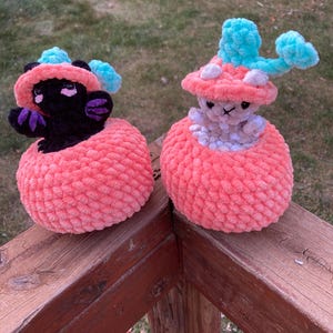 Puede incluir: Dos juguetes de peluche de crochet con forma de hongo con un pequeño animal asomándose por la parte superior. Los hongos son de color rosa y los animales son de color negro y blanco.