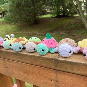 May include: A collection of colorful, crocheted turtle toys in various colors, including pink, purple, yellow, and green. The turtles are arranged on a wooden surface, with a natural outdoor background.