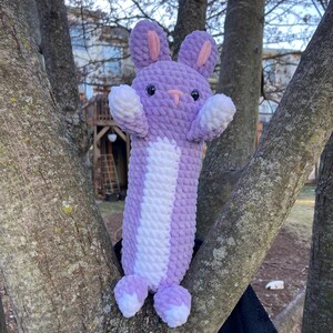 May include: A purple and white crocheted bunny toy with long ears and a long body. The toy is hanging from a tree branch.