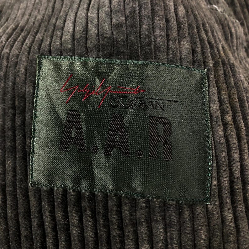 May include: Close-up of a dark gray corduroy fabric with a rectangular green label. The label features the text "Yohji Yamamoto DURBAN A.A.R" in red and white lettering. The corduroy has vertical ridges.