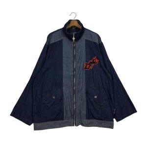 May include: A denim jacket with a zip-up front, featuring a colour-blocked design with a lighter grey panel down the centre. The jacket has a red and black embroidered logo on the chest and two front pockets.