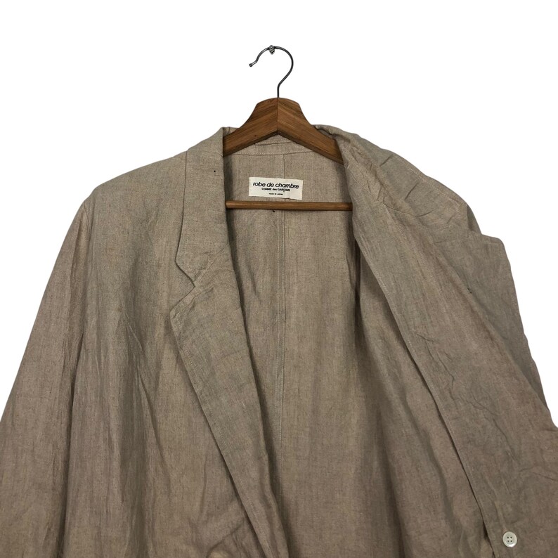 K&ouml;nnte beinhalten: Ein hellbeiger Blazer aus Leinen mit einem einzigen Knopfverschluss. Der Blazer hat ein Etikett mit der Aufschrift "robe de chambre" in der Mitte der Vorderseite.
