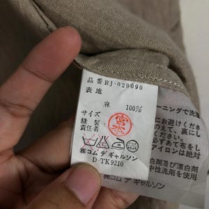 K&ouml;nnte beinhalten: Ein wei&szlig;es Etikett mit schwarzem Text und einem roten Stempel. Das Etikett ist an einem beigefarbenen Stoffkleidungsst&uuml;ck befestigt. Das Etikett lautet "品番 RJ-020090 表地 麻 100% サイズ 縫製 責任者 (株)コムデギャルソン D-TK 9210 ーニングで洗濯 にお避けくださ のえて、裏にし ください。 必ずあて布をし アイロンは絶対 白剤及び漂白剤 中性洗剤を使用 ㈱コムデギャルソン".