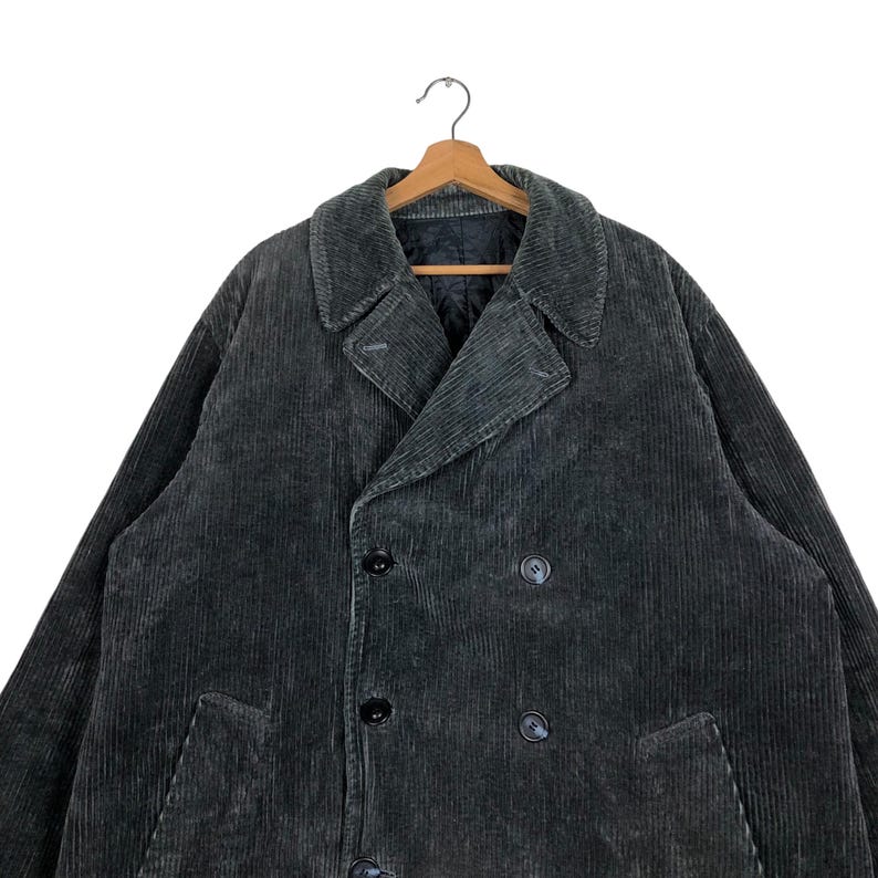 May include: A dark gray corduroy coat with a double-breasted design, featuring large lapels and black buttons. The coat is hanging on a wooden hanger against a white background. The fabric has a ribbed texture.
