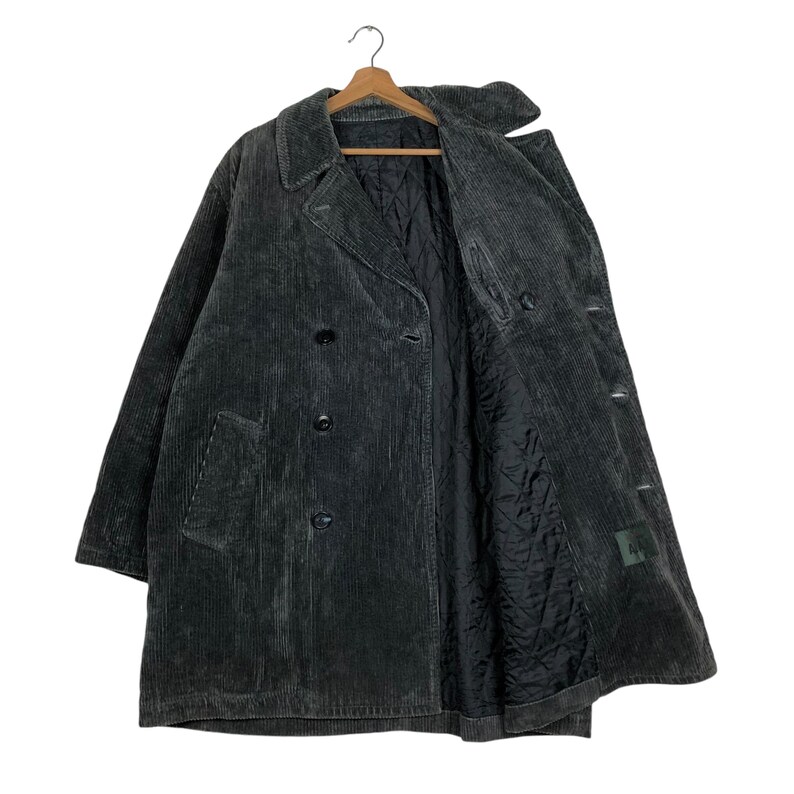 May include: A dark gray corduroy coat with a double-breasted design. The coat features a notched collar, two front pockets, and a quilted black lining. The coat is hanging on a wooden hanger.