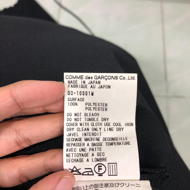 Puede incluir: Una etiqueta de prenda negra con texto blanco de COMME des GAR&Ccedil;ONS Co., Ltd. enumera los detalles de la tela y las instrucciones de cuidado. La etiqueta dice "MADE IN JAPAN" y "FABRIQUE AU JAPON". La tela es 100% poli&eacute;ster. La etiqueta tambi&eacute;n incluye s&iacute;mbolos de lavado.