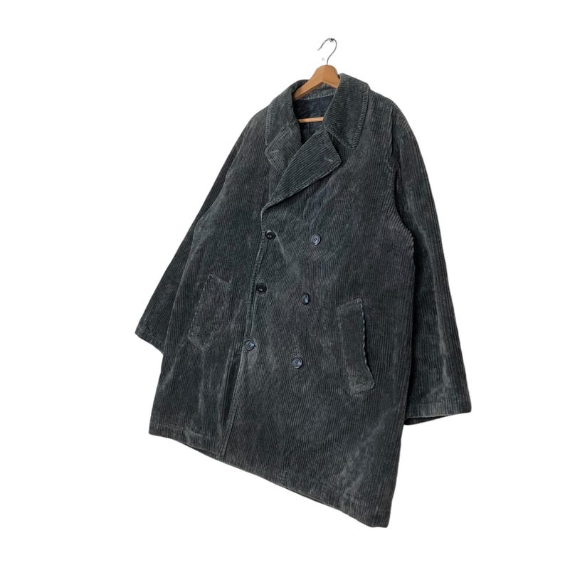 May include: A dark gray corduroy coat with a double-breasted design. The coat has a notched collar, two front pockets, and a button closure. The coat is hanging on a wooden hanger.