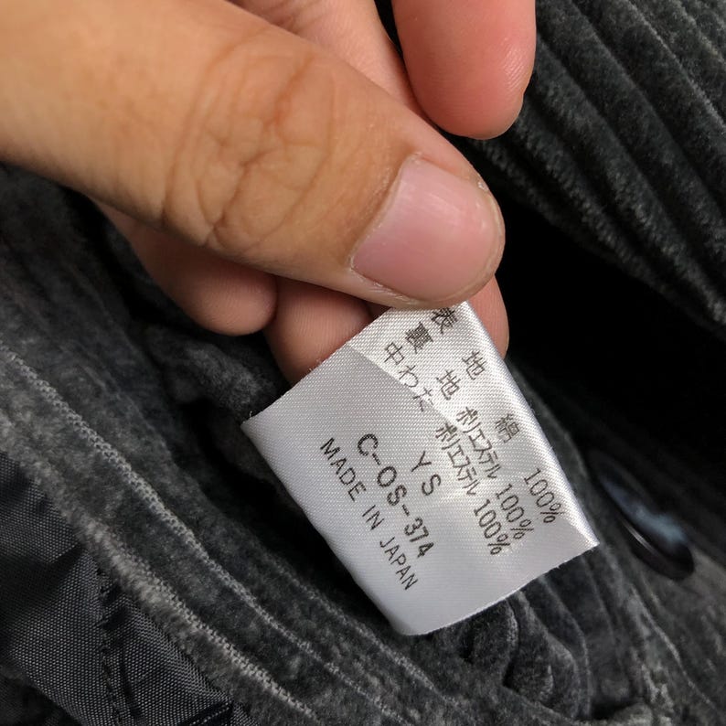 May include: A close-up of a white tag with Japanese text and the text "MADE IN JAPAN". The tag is held by a hand and is attached to a gray, corduroy-like fabric. The tag also has the text "C-OS-374".