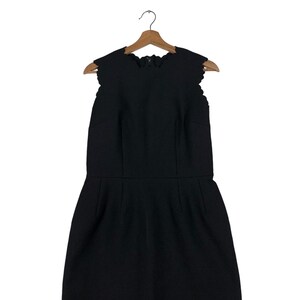 Puede incluir: Vestido negro sin mangas con escote y dobladillo festoneados, colgado de una percha de madera. El vestido tiene un corpi&ntilde;o ajustado y una falda ligeramente acampanada, creando una silueta cl&aacute;sica.