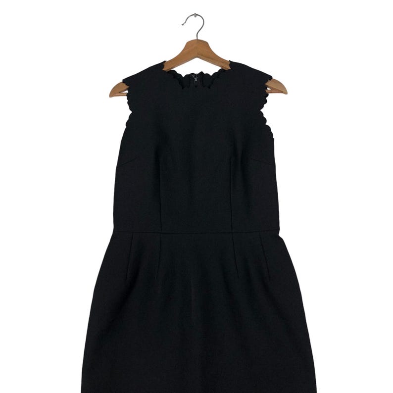 Puede incluir: Vestido negro sin mangas con escote y dobladillo festoneados, colgado de una percha de madera. El vestido tiene un corpi&ntilde;o ajustado y una falda ligeramente acampanada, creando una silueta cl&aacute;sica.