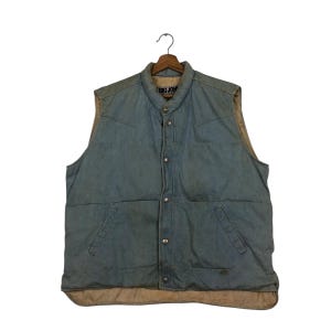 May include: A light blue denim vest with a quilted design. The vest features a button-up front, two front pockets, and a beige lining. The brand name "BIG JOHN" is visible on the collar.