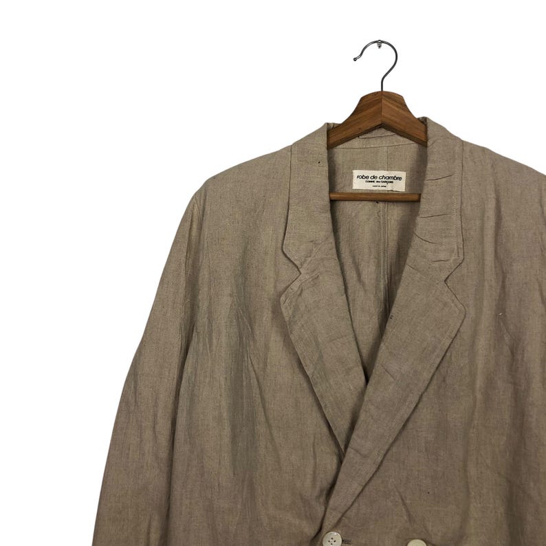 K&ouml;nnte beinhalten: Ein beiger Blazer aus Leinen mit doppelreihigem Verschluss und einem Etikett mit der Aufschrift "robe de chambre" am inneren Kragen.