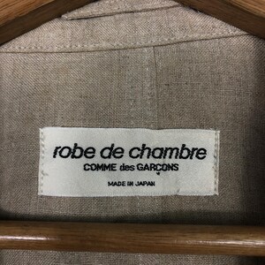K&ouml;nnte beinhalten: Eine beige Leinen-Hausjacke von Comme des Gar&ccedil;ons mit einem wei&szlig;en Etikett, auf dem "robe de chambre Comme des Gar&ccedil;ons Made in Japan" steht.