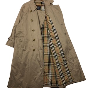May include: A tan trench coat with a double-breasted design, featuring a classic collar and button closures. The coat's interior lining showcases a plaid pattern in beige, red, and black. The coat is hanging on a wooden hanger.
