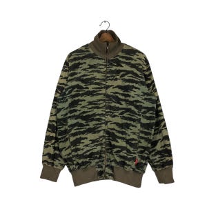 Puede incluir: Una chaqueta de manga larga con cremallera que presenta un patrón de camuflaje de rayas de tigre en verde y negro. La prenda incluye un cuello alto, puños acanalados y un dobladillo acanalado en color verde oliva sólido, con una etiqueta roja.