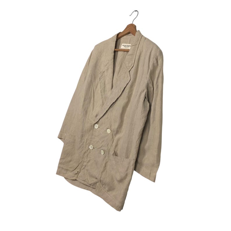 K&ouml;nnte beinhalten: Ein beiger Blazer aus Leinen mit doppelreihigem Verschluss und einer aufgesetzten Tasche auf der linken Seite. Der Blazer hat eine lockere Passform und eine lange, flie&szlig;ende Silhouette.