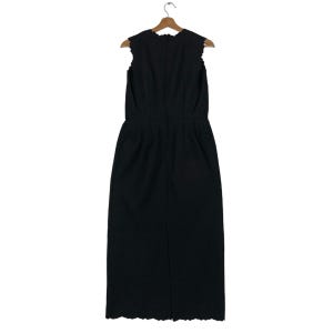 Puede incluir: Vestido negro sin mangas con escote y dobladillo festoneados, colgado de una percha de madera. El vestido tiene un corpi&ntilde;o entallado y una falda recta. La parte trasera del vestido presenta una costura vertical y una abertura en la parte inferior.