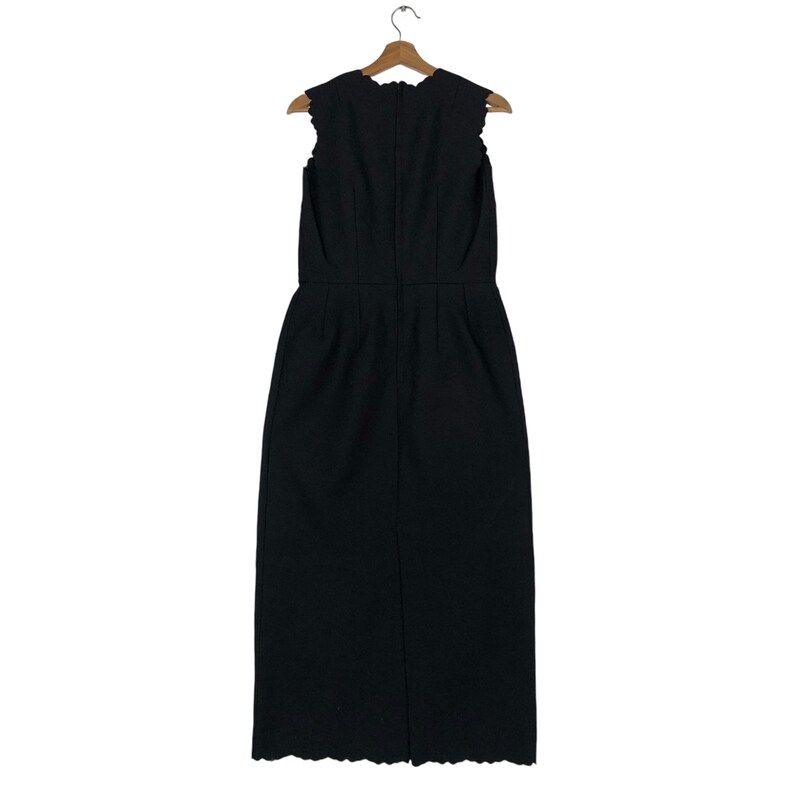 Puede incluir: Vestido negro sin mangas con escote y dobladillo festoneados, colgado de una percha de madera. El vestido tiene un corpi&ntilde;o entallado y una falda recta. La parte trasera del vestido presenta una costura vertical y una abertura en la parte inferior.