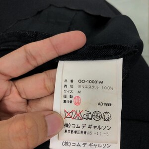Puede incluir: Primer plano de una etiqueta de prenda negra con texto japon&eacute;s y s&iacute;mbolos de cuidado. La etiqueta incluye el c&oacute;digo de producto GO-10001M, la composici&oacute;n del material, la talla M y el a&ntilde;o AD1998. La etiqueta est&aacute; adherida a una tela negra.