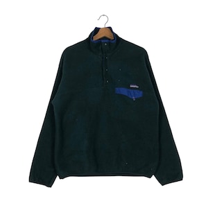 May include: A dark green fleece pullover with a blue pocket and collar trim. The long-sleeved top has a button-up closure and a small brand logo on the pocket. The garment is hanging on a hanger.
