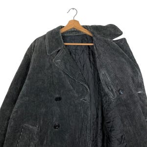 May include: A dark gray corduroy coat with a double-breasted design. The coat features a notched collar, two rows of buttons, and two front pockets. The interior lining is black and quilted. The coat is hanging on a wooden hanger.