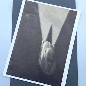 May include: Black and white photograph of a streamlined vehicle, possibly a race car or aircraft, with a long, pointed nose. The image is printed on a white-bordered card, resting on a dark gray background.