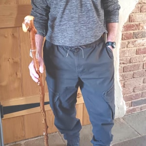 May include: A person wearing a grey zip-up sweater and dark grey cargo trousers. They are holding a wooden walking stick with a decorative handle. They are wearing black boots. The background includes a wooden door and a brick wall.