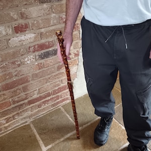 May include: A brown wooden walking stick with a decorative handle. The stick is held by a person. The walking stick is approximately 91 cm long.