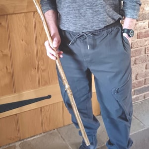 May include: A handcrafted wooden walking stick with a curved handle. The stick is light brown with a textured surface and a label attached. The person is wearing a gray shirt and dark pants.