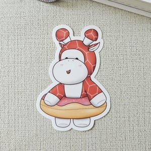 May include: A sticker featuring a cartoon giraffe wearing a red and white giraffe-patterned outfit and a pink donut float. The sticker is on a light gray surface, with a notebook and pen in the background.