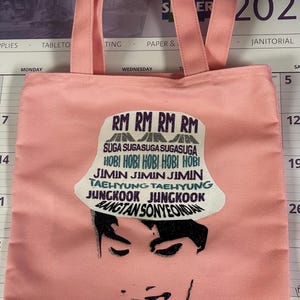 May include: Pink tote bag with matching handles. The bag features a graphic of a person's face and a hat with text that reads "RM, Suga, Hobi, Jimin, Taehyung, Jungkook, Bangtan Sonyeondan."