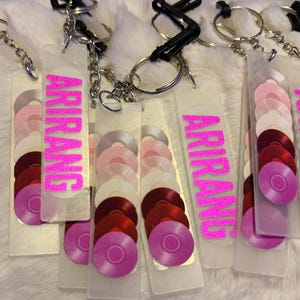 May include: Clear acrylic keychains with the name "ARIRANG" in bright pink glitter. Each keychain features a gradient of small, colorful vinyl record designs in shades of pink, red, and white, attached to a silver keyring.