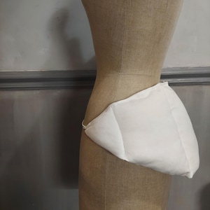 May include: A white fabric bustle pad worn around the waist, featuring a structured, padded design that extends outward from the back. The garment is made of smooth, plain white material and is secured with a thin strap around the torso.