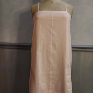 Puede incluir: Un camisón rosa claro con tirantes finos y un escote recto. El vestido se exhibe en un maniquí vintage. El escote presenta un delicado detalle bordado. La tela parece ligera y fluida.