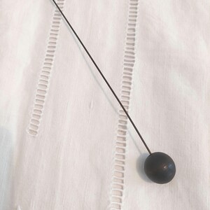 May include: A dark metal hat pin with a round, ball-shaped head. The pin is long and slender, with a sharp point. The pin is set against a white background with decorative stitching.