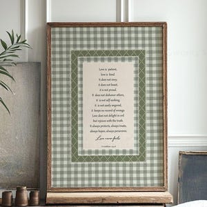 Puede incluir: Una impresión de arte mural enmarcada con un borde de patrón de cuadros vichy en verde salvia y blanco que rodea un panel central color crema. El texto muestra el pasaje bíblico 1 Corintios 13:4-8, terminando con la frase Love never fails.