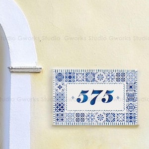 Puede incluir: Placa rectangular con número de casa, fondo blanco y borde decorativo azul. El número "575" se muestra en azul degradado. La placa está montada en una pared de color amarillo claro.