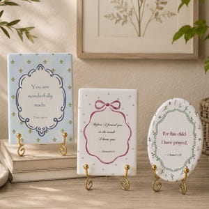 May include: Three decorative ceramic plaques with inspirational quotes. The plaques are rectangular and oval, featuring floral and ornate designs. Each plaque is displayed on a gold-coloured metal stand. The text includes phrases like "You are wonderfully made" and "For this child I have prayed."