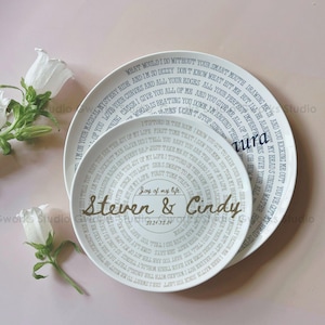 May include: Two white ceramic plates with a circular design featuring song lyrics and names. The plates have a light grey text and a gold-coloured text that reads "Steven & Cindy" and the date "2024.12.04".