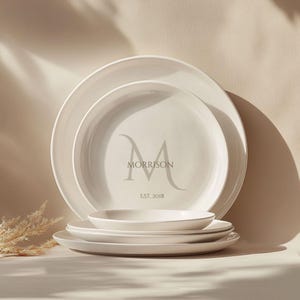 Puede incluir: Un conjunto de platos y cuencos de cerámica blanca. El plato más grande tiene un monograma gris con el nombre "MORRISON" y el año "EST. 2018". Los platos están apilados, con un cuenco pequeño encima.