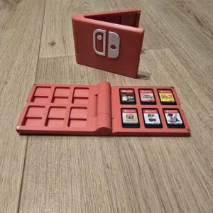 May include: A coral-coloured Nintendo Switch game cartridge holder, shaped like a wallet, with the Switch logo on the front. The open holder displays game cartridges. The holder is on a wooden surface.