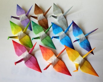 May include: A collection of ten origami cranes folded from colourful paper. The cranes are arranged in a circle, with the tips of their wings pointing outwards. The colours of the cranes are purple, yellow, green, blue, orange, brown, and grey.