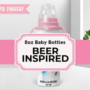May include: A white 8oz baby bottle with a pink handle and nipple. The bottle has the text "BEER INSPIRED" and "BORN IN THE ROCKIES EST. 1978". The bottle is designed to look like a beer bottle.