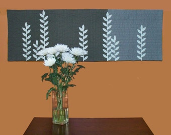 May include: A white vase with white flowers sits on a dark brown table in front of a wall with a gray and white patterned fabric hanging on it.