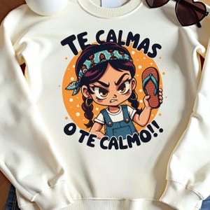 May include: White sweatshirt featuring a cartoon illustration of a girl with brown hair wearing a blue and white headband and a blue overall dress. The girl is holding a blue flip-flop and has an angry expression. The text "TE CALMAS O TE CALMO!!" is printed on the sweatshirt.