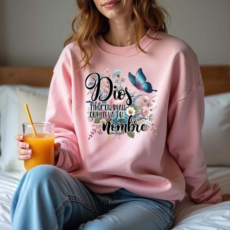 May include: Pink sweatshirt with a floral design and a butterfly. The text on the sweatshirt reads "Dios tiene un plan que lleva tu nombre".