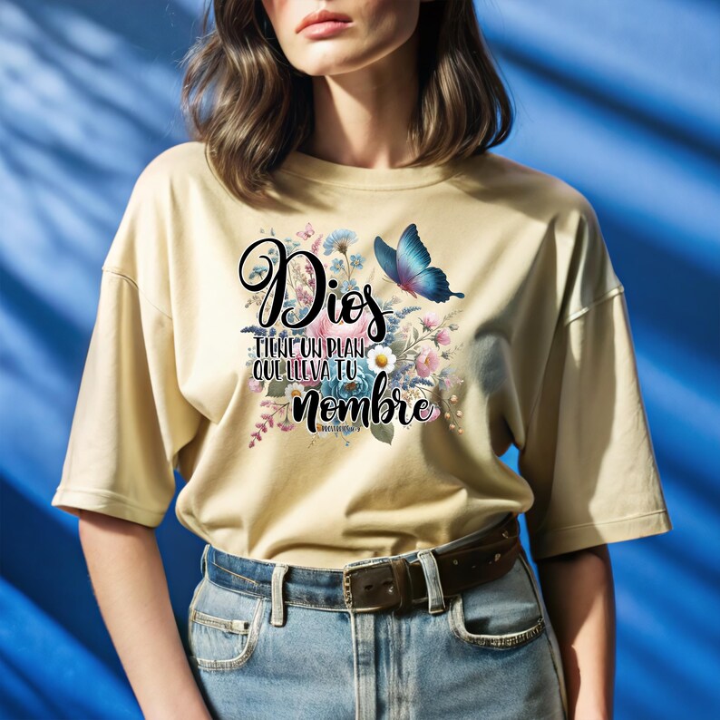 May include: A beige t-shirt with a floral design and a butterfly. The text on the shirt reads "Dios tiene un plan que lleva tu nombre" with a scripture reference below.